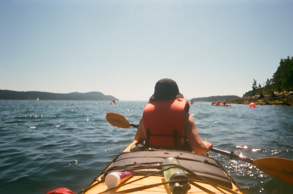 Young person wearing a black hat and lifejacket sits in a kayak facing the mountains and holding a paddle in front of them.