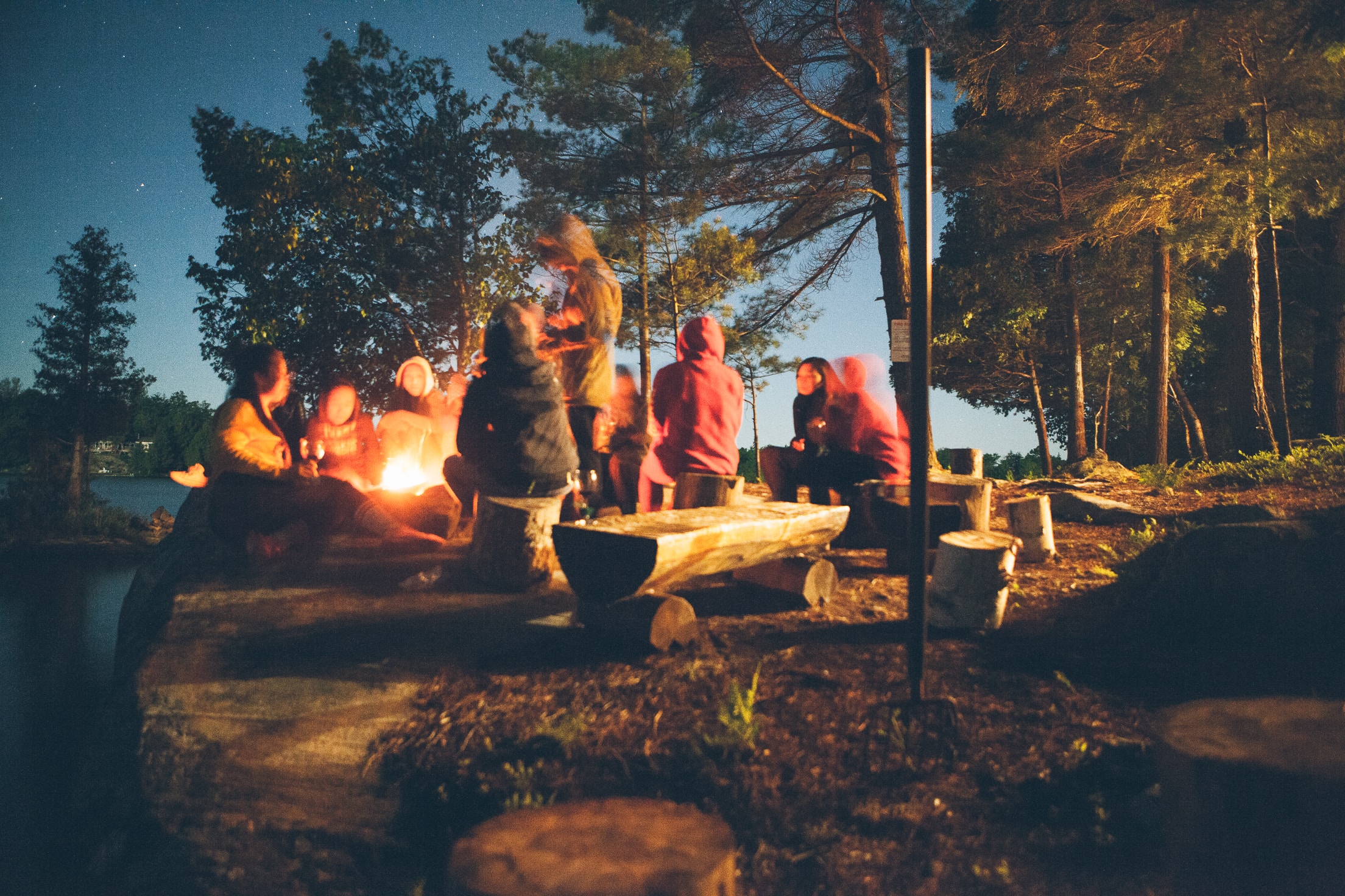 8 people wearing sweaters are getting cozy around a campfire at dusk