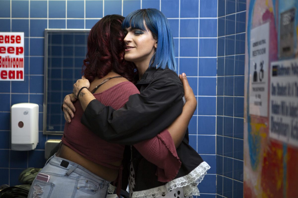 A non-binary femme embracing another student in a school bathroom.