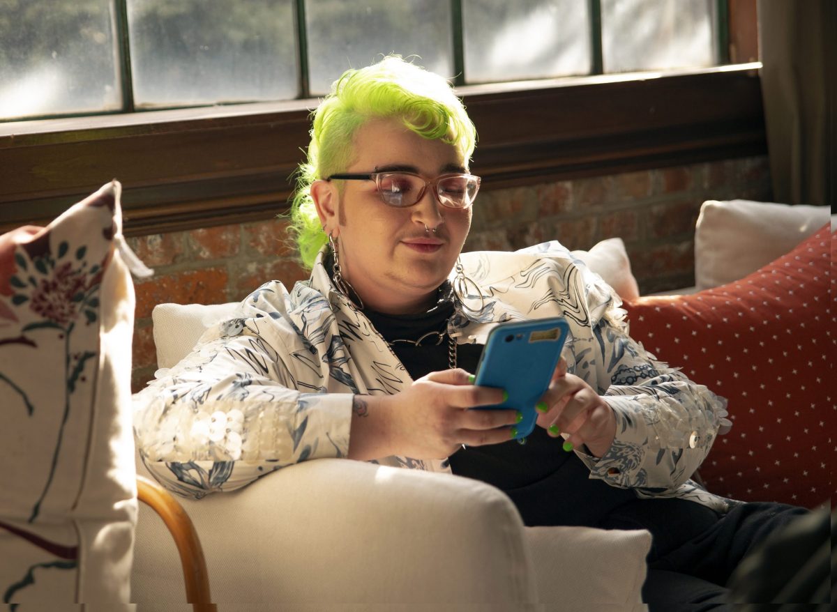 A genderqueer person sitting on a couch looking at their phone