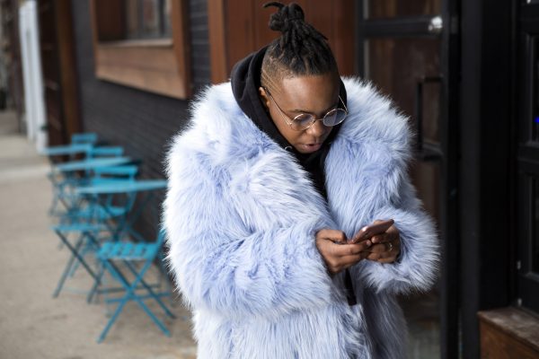 A transmasculine person with a furry blue coat checking his phone on the sidewalk scaled.