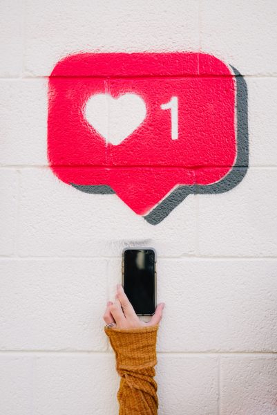 Arm wearing an orange sweater holds a black phone against a white wall, an instagram "like" icon is painted on the wall above the phone